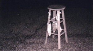 Black and white photograph of a tall wooden stool with a pair of satin ballet slippers tied to the front legs, standing in the corner of a carpeted room.