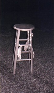 Black and white photograph of a tall wooden stool with a pair of satin ballet slippers tied to the front legs, standing in the corner of a carpeted room.