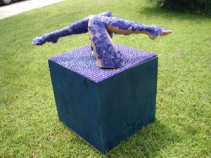 Outdoor sculpture of two human legs covered in textured blue material emerging from the top of a cube covered in purple pegs, placed on grass near trees.