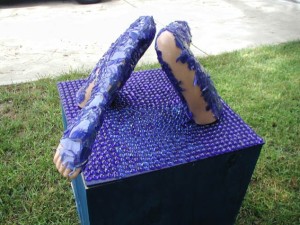 Outdoor sculpture of two human legs covered in textured blue material emerging from the top of a cube covered in purple pegs, placed on grass near trees.