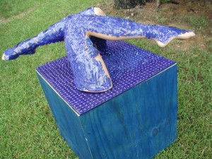 Outdoor sculpture of two human legs covered in textured blue material emerging from the top of a cube covered in purple pegs, placed on grass near trees.