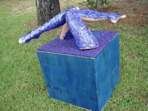 Outdoor sculpture of two human legs covered in textured blue material emerging from the top of a cube covered in purple pegs, placed on grass near trees.