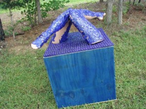 Outdoor sculpture of two human legs covered in textured blue material emerging from the top of a cube covered in purple pegs, placed on grass near trees.