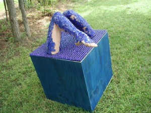 Outdoor sculpture of two human legs covered in textured blue material emerging from the top of a cube covered in purple pegs, placed on grass near trees.