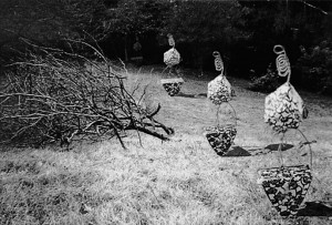 Outdoor installation of humanoid sculptures made of patterned paper and wire, placed in a row on a grassy slope near a fallen tree.