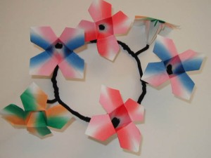 Wall-mounted wreath-like structure made of origami flowers in pink, blue, green, and white, connected by black pipe cleaners.