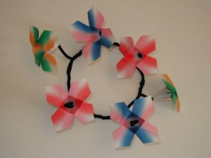Wall-mounted wreath-like structure made of origami flowers in pink, blue, green, and white, connected by black pipe cleaners.