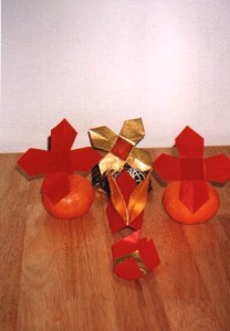 Origami flowers in red and gold paper, some mounted on oranges, arranged on a wooden table with a white background.